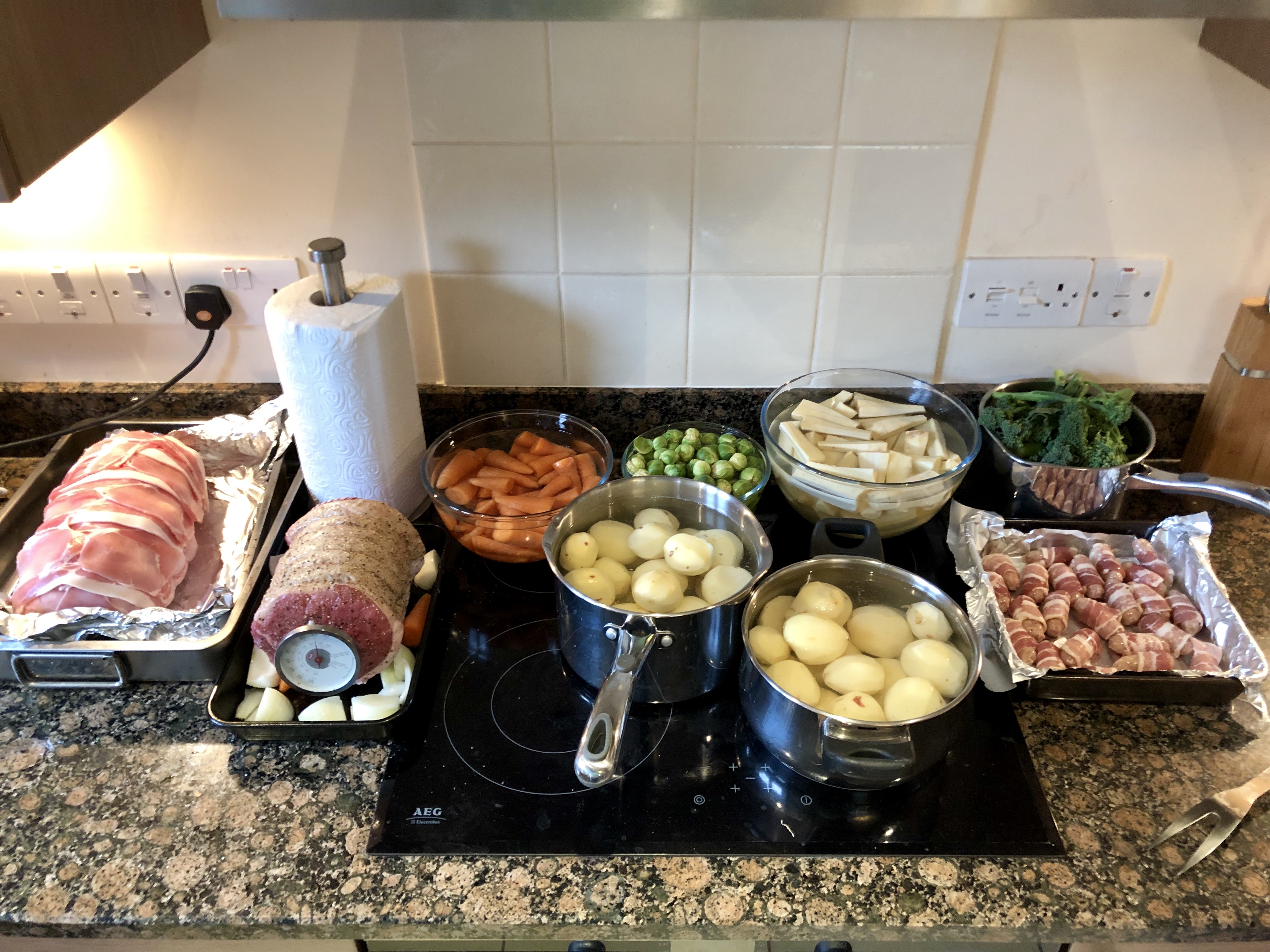 The great Christmas dinner thread - Page 3 - Food, Drink & Restaurants - PistonHeads - The image shows a kitchen countertop laden with various types of food. In the foreground, there are pots and pans filled with different ingredients. There's a large roast in one pot, and it appears to be partially covered by foil. Beside the roast, there are several bowls containing additional dishes; one bowl seems to contain vegetables while another contains potatoes. Additionally, there is a tray of meat and some spoons on the countertop. The kitchen has a white tiled floor and cabinets with brown doors. A light hangs from the ceiling over the counter, casting light onto the food. There's no visible text in the image.