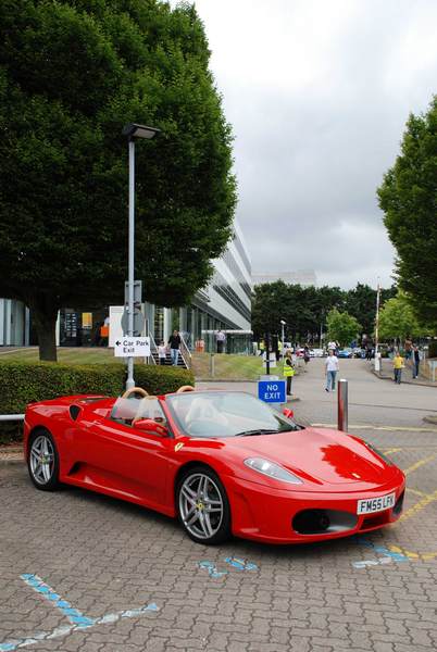 Service Sunday Pistonheads - This image features a sleek red sports car parked in an outdoor lot. The car is facing towards the left side of the frame and has a distinctive design with a long hood and short rear deck, accentuated by large wheels and a low ground clearance. The vehicle is parked near the curb, with a sign appearing to be a "No Parking" zone in the foreground.

The sky in the background is cloudy, suggesting an overcast or possibly rainy day. The setting seems to be a streetside parking area adjacent to a building, with the car positioned to make a right turn onto the road. There are other cars and people parked or walking in the background, indicating an active urban environment. The lighting in the photo is soft, which could be a result of the light diffusing through the clouds.