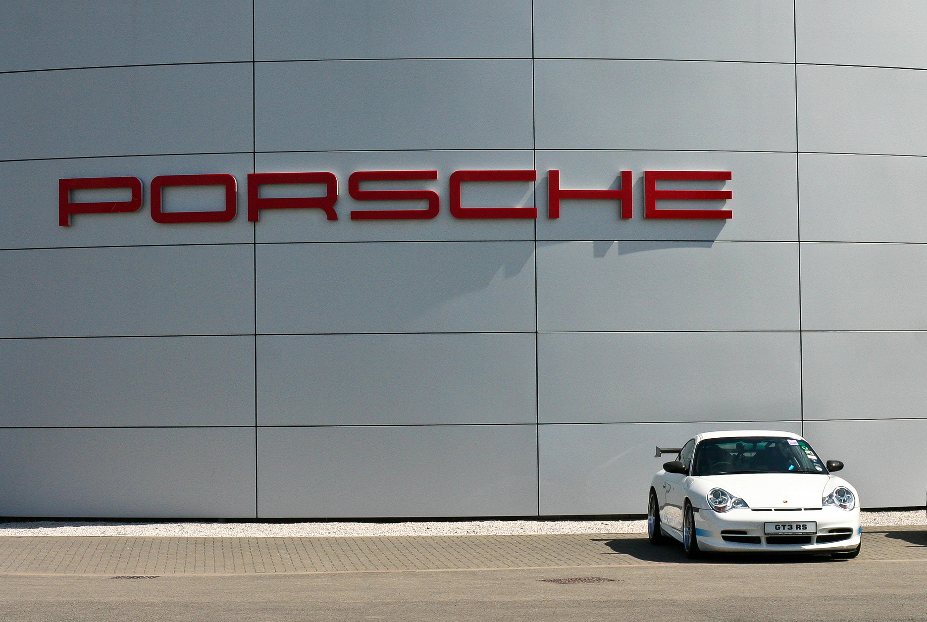 A white toilet sitting on top of a street - Pistonheads - The image is a stylized photograph of a Porsche emblem located on the front of a large building. The building facade is composed of vertical lines that appear to be made from a shiny, reflective material. In front of the building, a small white sports car is parked. The car is angled diagonally against a backdrop of gravel. The overall composition of the image suggests a theme related to automotive design, perhaps at a car dealership or a car manufacturing facility.