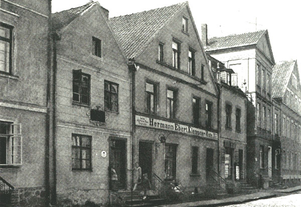 The image is a black and white photograph capturing an architectural scene. It depicts a row of old, one-story brick buildings with slanted roofs, each sitting atop its stone foundation. These buildings appear to be residential, characterized by numerous windows and simple doorways. A sidewalk is visible in front of the buildings, with some individuals visible as well. Above one of the doors, there is a business sign with the name "Hermann Erbstein," indicating a commercial component to the architecture, suggesting that this was once a main street in a European city. The clear sky above suggests it could be a sunny day.