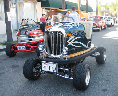 Dodgems Pistonheads Recycling - The image features a street scene with several vintage and custom vehicles parked. The most prominent vehicle is a modified car with a streamlined body, having been converted to a hot rod with a wide stance, raised front end, and a drag racing style engine cover. The car is painted in a combination of black and chrome accents with heat-resistant upholstery inside, providing visible contrast between the interior and exterior.

Additionally, other vehicles and pedestrians in the background suggest a public location, possibly a car show or an event where enthusiasts gather to display their designs and modifications. The tableau showcases a vibrant community engaged in the passion of vintage and performance automobiles. The vehicles capture attention and reflection of the popular trends in automotive design from several decades ago.