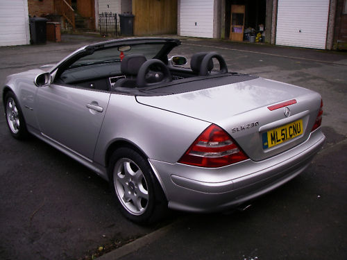 Show us your Mercedes! - Page 8 - Mercedes - PistonHeads - The image displays a sleek gray convertible car parked on a paved surface. The vehicle is featuring the convertible top open, revealing two empty seats inside, and the driver's side open, allowing a view into the cabin. A yellow license plate is discernible on the back of the car, indicating its registration. The setting appears to be a residential area with houses visible in the background, and the ground shows signs of recent rain, making it wet and reflective.