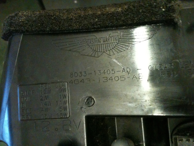 Db9 rear brake light - Page 1 - Aston Martin - PistonHeads - The image shows a piece of machinery with a brushed metal surface. There's a rectangular stamp in the center with alphanumeric characters, which is likely to be the model number or serial number, though the specific details are not clear. The machine has a black rubber component at the top, and there are two screws, one on each side of the metal body. The background is a nondescript grey, providing a neutral backdrop that highlights the machinery.