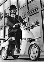 A 'period' classics pictures thread (Mk III) - Page 16 - Classic Cars and Yesterday's Heroes - PistonHeads UK - The image is a black and white photograph featuring a man riding a small moped. He appears to be in mid-motion, with one foot on the pedal and the other hand on the handlebar. His attire consists of a suit jacket and tie, and he has a mustache and beard. In the background, there is a brick wall and a large window that suggests an urban setting, possibly a building. The man's pose and the setting suggest this could be a candid or historical photograph.