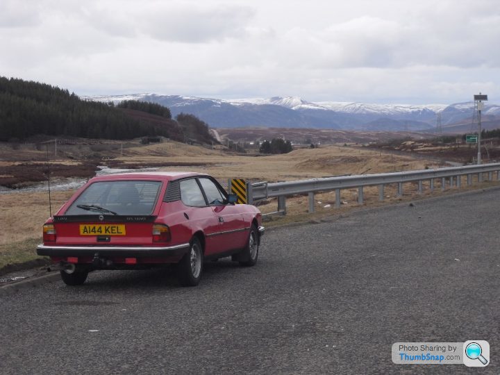 A car is parked on the side of the road - Pistonheads - The image shows a red car parked on the side of a highway. It seems to be a calm day as the car is parked in a serene setting. The highway itself is clear of vehicles and is bordered by a metal railing on one side and a green field on the other. The landscape includes mountains and a large body of water. The key hairpin curve of the highway is coming up ahead.