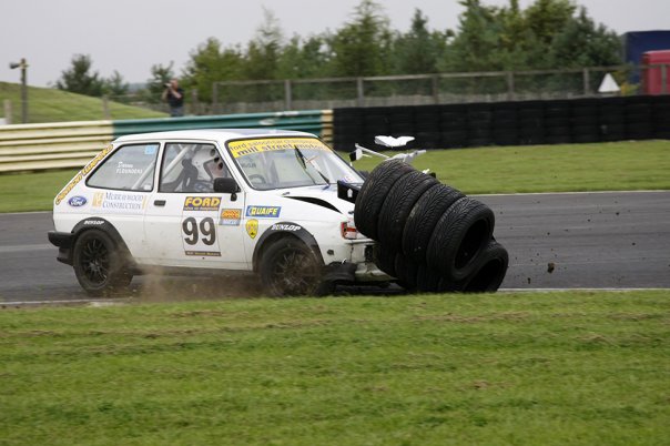 croft --- 26 & 27 sept   anybody ? - Page 1 - North East - PistonHeads - The image shows a scene of a white car that is positioned in a grassy area next to a track. The car appears to be a vintage Ford rally car with the number 99 on it, suggesting participation in a rally or racing event. It has a significant amount of tire tread left and is seen swaying and tipped over to the side. The interior of the car is exposed, and the driver's cockpit area can be seen. In the background, there's a person standing near the track, possibly observing the aftermath of the incident. The setting is an outdoor racing venue or course, as indicated by the tracks in the background and the grassy infield area where the car is located.