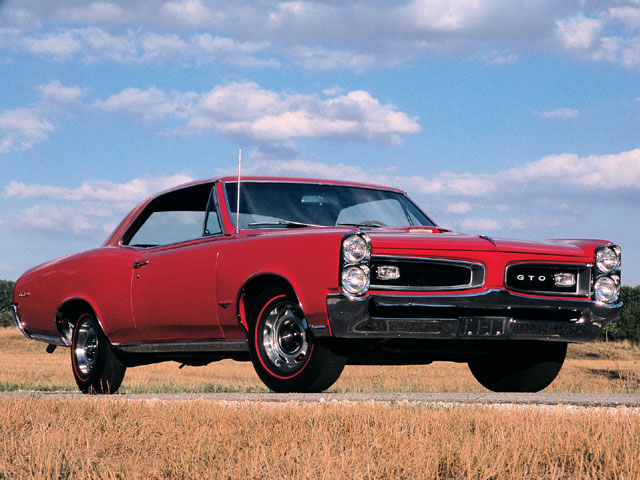 71 GTO in classifieds, what has it not sold? - Page 1 - Yank Motors - PistonHeads - The image features a classic-style classic red car, likely a Pontiac GTO, with a prominent white stripe running along its hood. The car is situated on a country road lined with tall golden grass. The sky in the background is a clear blue with a few scattered clouds. The setting appears to be on a rural road during the day.