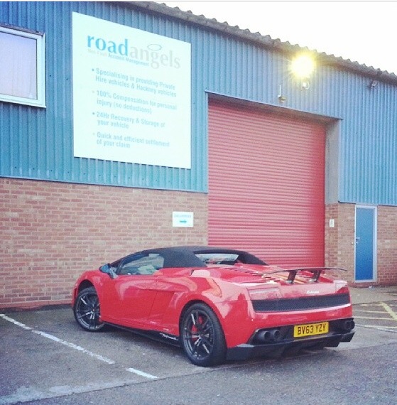 Performante, colours, numbers etc - Page 7 - Gallardo/Huracan - PistonHeads - The image shows a vibrant red sports car parked in front of a blue building. The vehicle has a prominent rear wing and a sleek design. There's a text sign on the side of the building with various statements, including business hours and offers for bodywork and vehicle maintenance. Additionally, there is a security light above the door of the building. The setting appears to be a commercial or industrial area, indicated by the presence of a garage door.