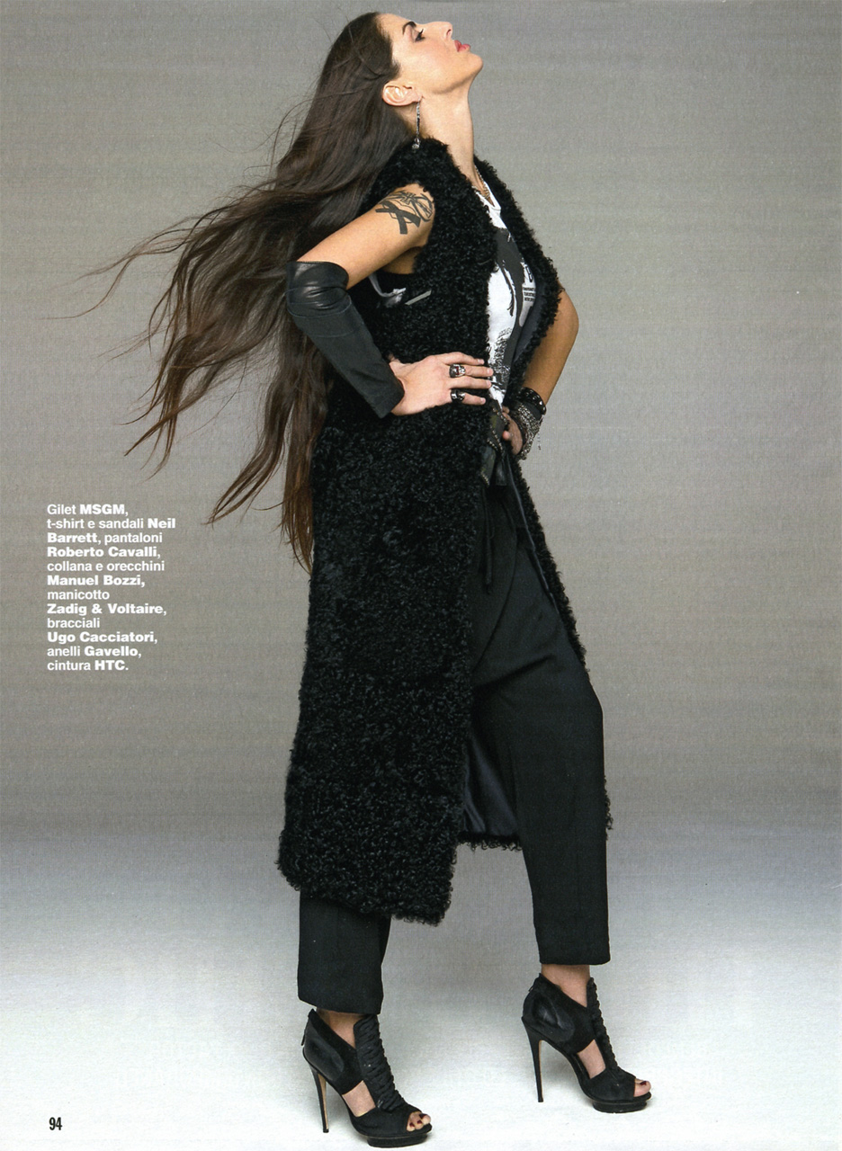 The image features a stylish individual posing and looking upwards and to the side. They have long hair that flows slightly down. They are wearing a black, fluffy coat and black pants. The coat has a distinctive fur design on the hood. The person is adorned with red bracelets and has a ring on one finger. They are wearing black high-heeled shoes with straps, complementing their outfit. The pose suggests confidence and strength.