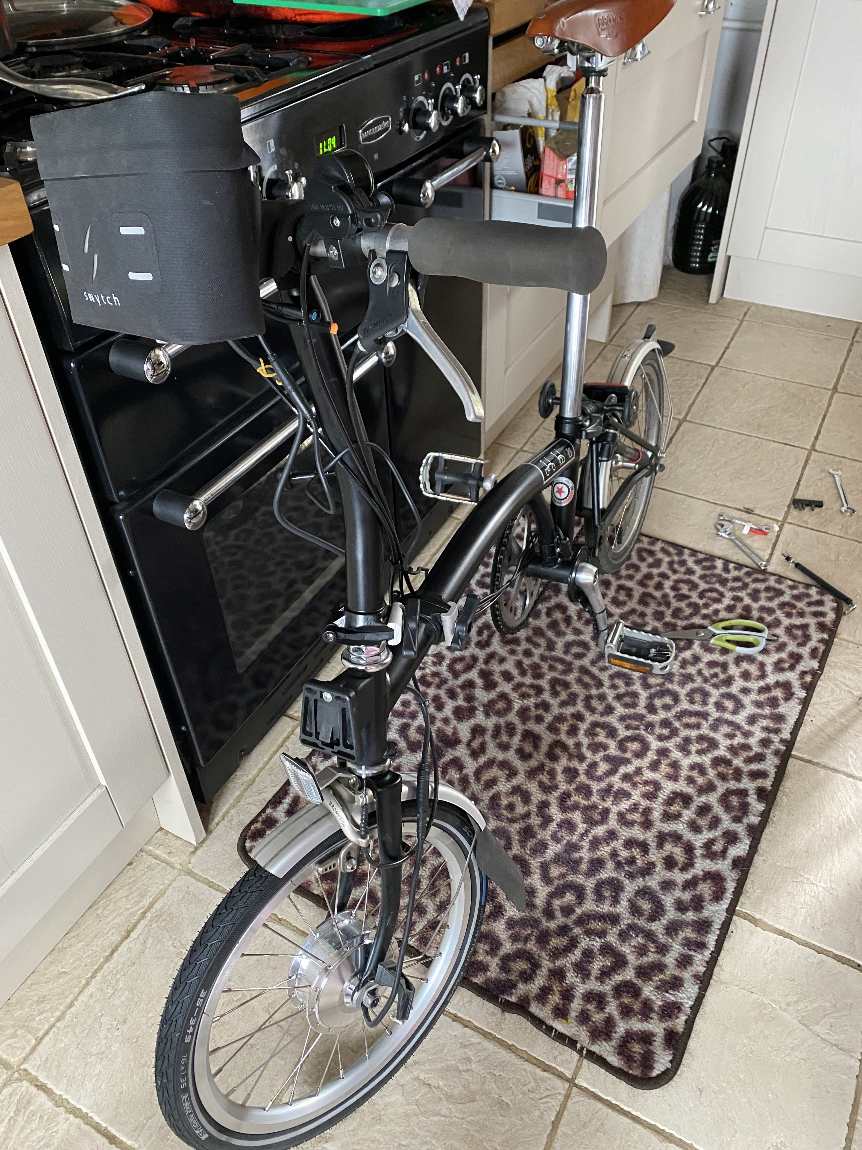 Let's see your Brompton  - Page 22 - Pedal Powered - PistonHeads UK - The image shows a black folding bicycle set up on a kitchen floor. The bicycle is unfolded and leaning against the wall, with its seat and handlebars at an angle, indicating it's ready to be used or stored. There are various items around the bicycle, including what appears to be a small appliance in the background and a kitchen table. The scene gives a homely vibe, suggesting that this might be someone's personal space where they keep their folding bicycle when not in use.