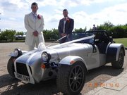 Soon be a 7 owner and just saying hi. - Page 1 - Caterham - PistonHeads - The image shows two men standing next to a vintage silver open-top car, which appears to be a classic racing model. Both men are dressed formally in suits and ties, suggesting they are attending a formal event or a wedding. The car is parked on what looks like a sidewalk or pavement, and the background features vegetation and a clear sky with scattered clouds, indicating it might be a sunny day. The overall scene suggests a special occasion or celebration with a nostalgic vehicle as a focal point.