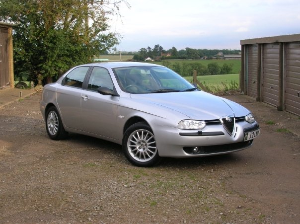 Pistonheads - The image features a silver Alfa Romeo car parked on a dirt road next to a metal shed. The car is positioned against the backdrop of the field and rolling hills, creating a contrast between the rural setting and the sleek design of the car. The dirt road suggests a rustic or rural environment, while the metal shed and the car hint at some level of human activity or even agriculture. The sky above is overcast, adding a dramatic touch to the scene.