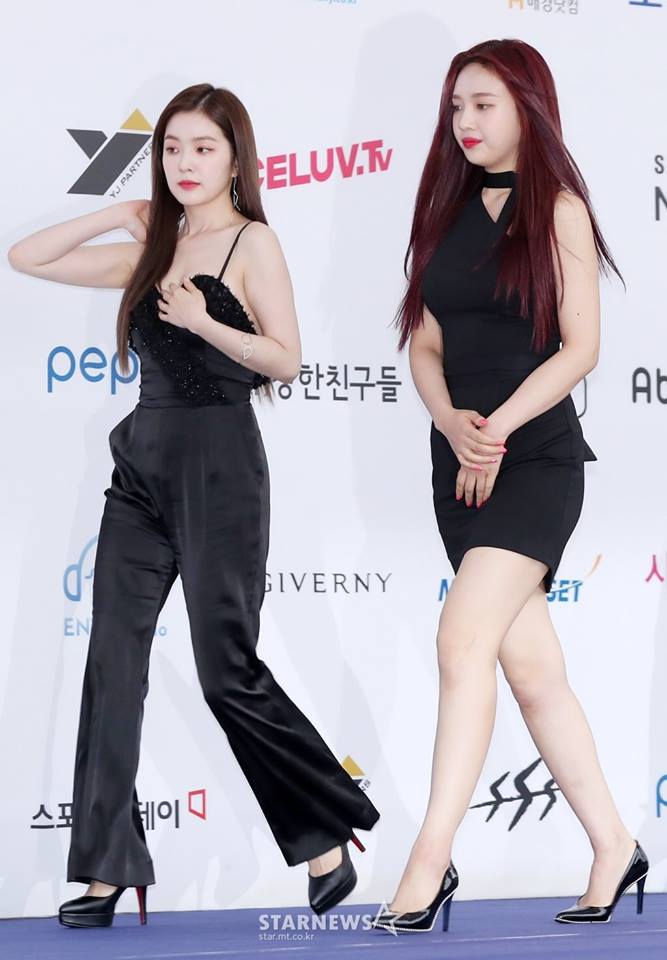 A woman and a woman standing next to each other - The image captures a moment on a red carpet, featuring two women walking side by side. The woman on the right stands out with her long, dark hair and black outfit, while the woman on the left sports a darker shade of hair and a similar black dress, her hand resting casually on her hip. They both appear confident and stylish, reflecting the glamorous vibe of the event they are attending. The red carpet stretches out behind them, leading to what looks like a stage with various logos and sponsors displayed.