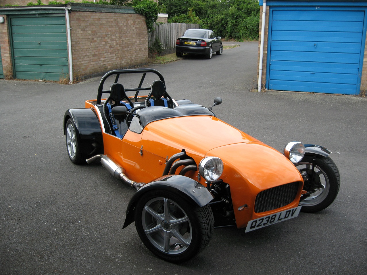 Pictures of your Kit Car..? - Page 36 - Kit Cars - PistonHeads - In the middle of the image, there's a vibrant orange and black convertible sports car with two seats, parked on a black asphalt road. The driveway appears to be empty except for the sports car, with the backdrop being a green garage and trees. A few leaves are visible on the ground, suggesting it might be autumn. The sports car is the focal point of the scene.