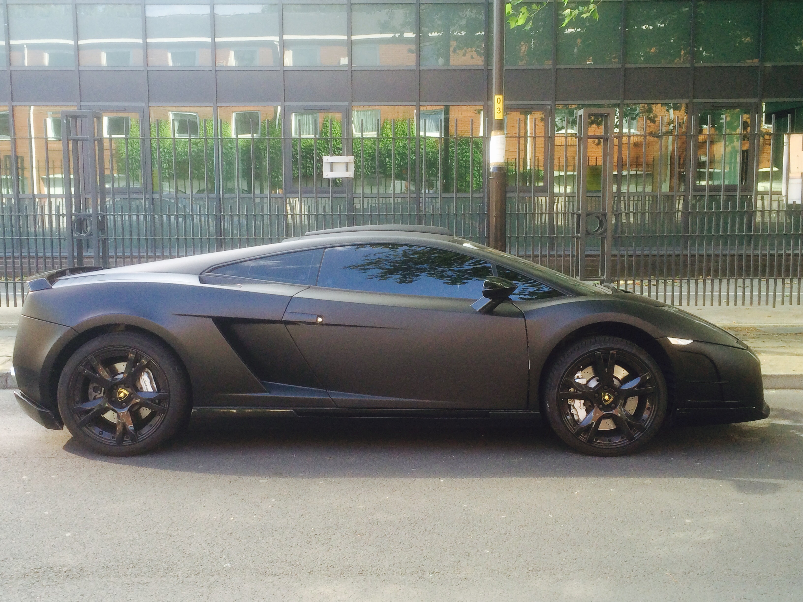 - Page 502 - North West - PistonHeads - The image shows a sleek, black sports car parked on the side of a road. The car's design features a prominent front spoiler, side mirrors, and air vents, which are indicative of its high-performance capabilities. The background consists of a building with large windows reflecting greenery, suggesting an urban setting with some level of greenery nearby. The image is taken during the day under clear skies, and the car's condition appears to be very clean and well-maintained.