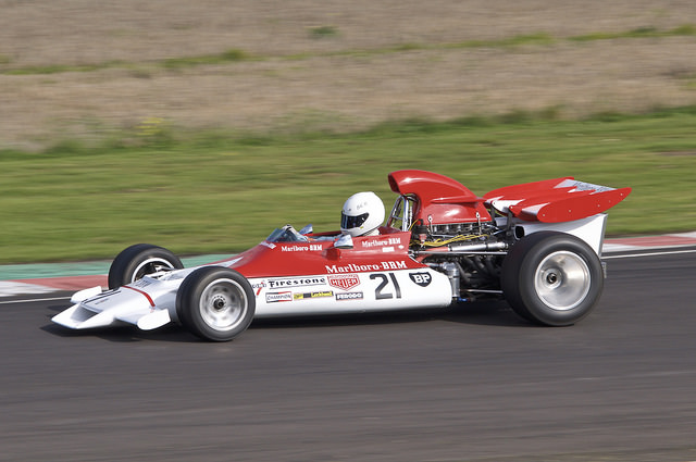 Photography at Castle Coombe - Page 1 - South West - PistonHeads - The image captures a vintage race car on a track, with a passionate driver at the wheel. The car is colorful, with a vibrant red and white livery. The action shots depict movement, with the driver maybe leaning into a corner. The car is labeled with numbers and sponsor names. The track and surroundings suggest an outdoor venue.