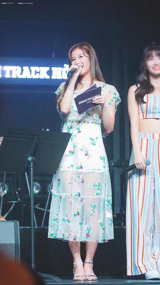 A woman holding a cell phone up to her ear - The image showcases two individuals on a stage with a microphone stand in the foreground. The person on the left is singing and holding a microphone with both hands, while the person on the right is standing slightly behind and to the side. The speaker is wearing a patterned, knee-length dress with lace details and is holding a bouquet of flowers. The overall ambiance suggests this could be a live performance or event.