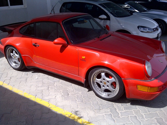 A car with a surfboard strapped to the back - Pistonheads - The image features a red Porsche 911 parked in what appears to be a parking lot. The car is positioned facing towards the left side of the frame, and its sleek design, characterized by the iconic shape and the details like the air intake above the recessed side vents, are clearly visible. 

The car is parked next to a silver sedan, indicating a well-used parking facility. The setting includes a concrete surface marked with a yellow stripe, which is likely indicating a designated parking space. There are no people or other objects immediately noticeable in the vicinity of the car. 

The image exudes a sense of elegance and exclusivity associated with Porsche vehicles. The contrast between the vibrant red of the Porsche and the more subdued colors of the car next to it further highlights the attention that the Porsche 911 attracts.