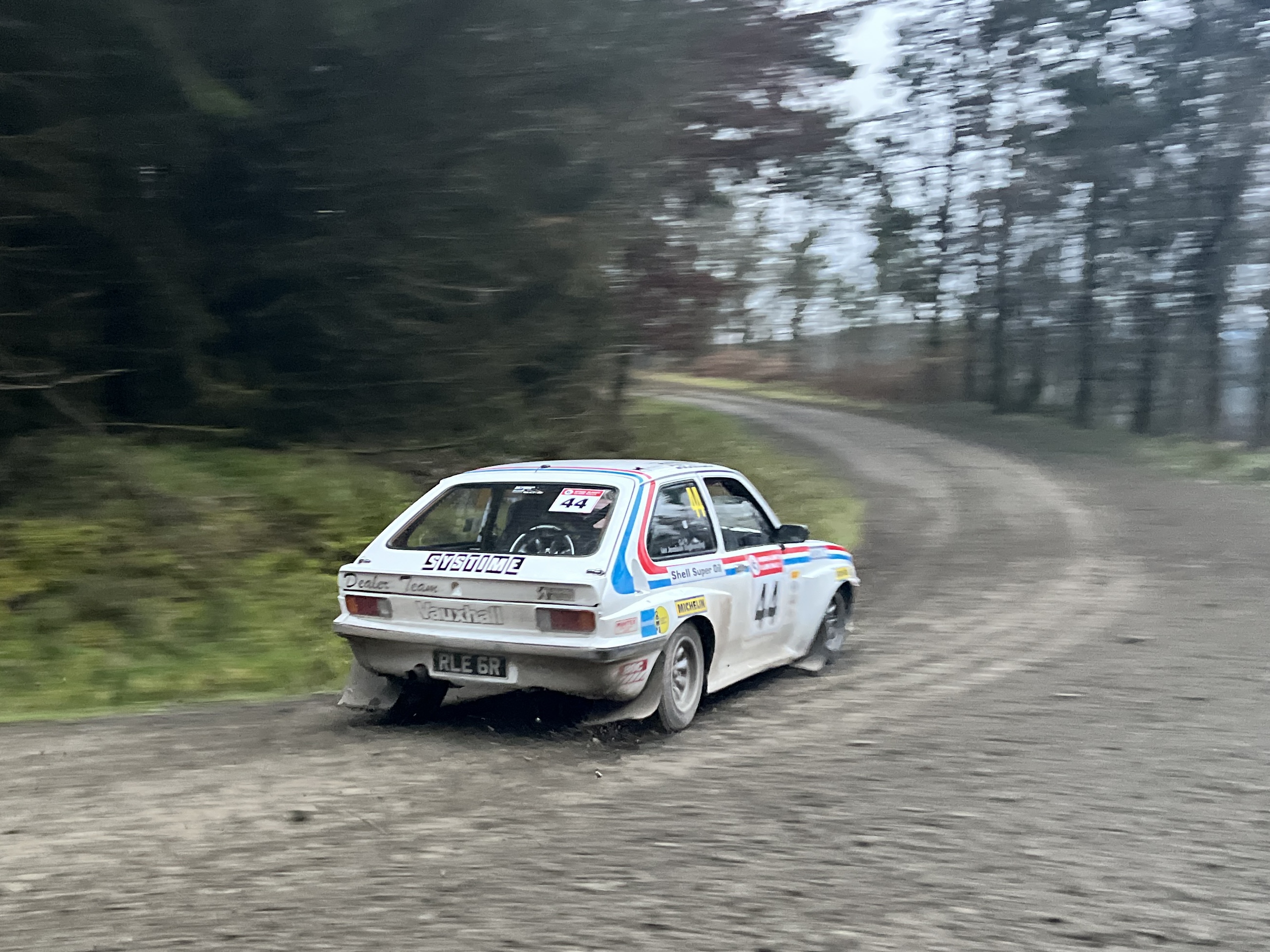 Roger Albert Clark Rally 2023 - Page 6 - Rallying - PistonHeads UK - The image features a vintage car in motion on a gravel road. The car is white with black markings and a red stripe along its side. It's an old-style rally car, likely from the 1970s or 1980s based on its design. There is a visible number "2" painted on the car, suggesting it might be participating in a racing event or rally stage. The setting appears to be a rural area with trees and grasses in the background. The style of the image is a real-life photograph that captures the movement and dynamism of the vehicle.