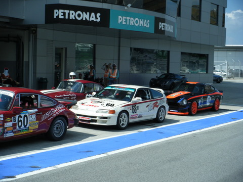 Sepang Afos Pistonheads - The image captures an outdoor scene with several vintage cars aligned side by side on a lot. The vehicles, which are the main focus of the image, exhibit various designs and colors, reflecting the diversity of the cars present. Some of the cars have parked, while one appears to be in motion, contributing to a dynamic composition. The lot is located in front of a building with large glass windows and a sign that reads "PETRONAS PETROMAS." A few individuals can be seen scattered around the scene, possibly car owners or spectators admiring the collection. The area is likely a gathering spot for enthusiasts of classic cars, with the cars situated on a road adjacent to the building, suggesting this may be a designated parking area or an event location.