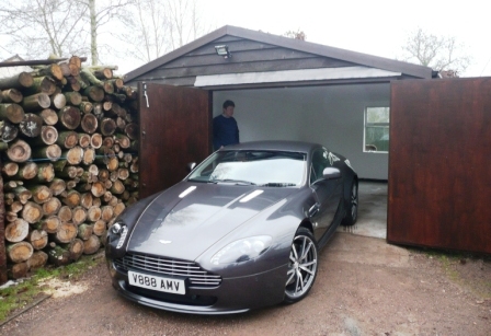 Delivery day - Page 1 - Aston Martin - PistonHeads - The image shows a man standing in a garage filled with wooden logs, behind a wooden door. The garage has a sloping roof and the man is dressed in casual clothing. A blue sports car is parked in the foreground, facing the viewer, with distinctive white stripes along the hood and side. There is a plate indicating "V8 500 AVG." The gravel driveway of the garage contributes to the rustic setting.