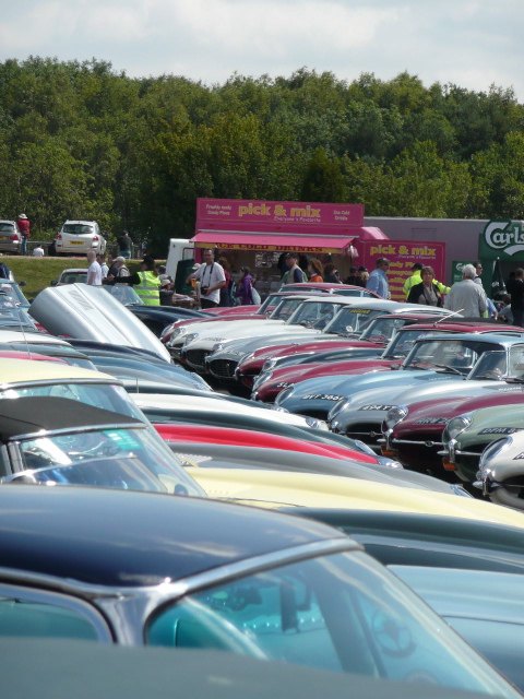 silverstone classic - Page 1 - Northamptonshire - PistonHeads - The image depicts an outdoor event with a parking lot filled with a variety of classic and vintage cars. Some cars are close to the camera, while others are further away, creating a layered effect of depth. The cars are primarily two-door models with a mix of soft-top and hard-top convertibles. There is a diversity of colors and model years, with a noticeable prevalence of traditional colors like red, black, and teal. A red and white fabric booth or tent can be seen behind some of the cars, indicating a likely car show or similar event in progress. The surrounding environment consists of vegetation and a building with prominent product advertising on the side. This setting suggests a community gathering focused on automobiles, likely during the day given the natural light.