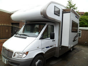 Selling RV? Best Way - Page 1 - Tents, Caravans & Motorhomes - PistonHeads - The image shows a parked recreational vehicle (RV). It has a light-colored exterior with a darker top and darker stripe, possibly on the side. The RV appears to be a van style conversion, and it is positioned on what looks like a paved surface. The background includes a brick wall and a portion of a tree, suggesting the RV is parked in an urban or suburban area. There are no visible texts or distinctive brands in the image.