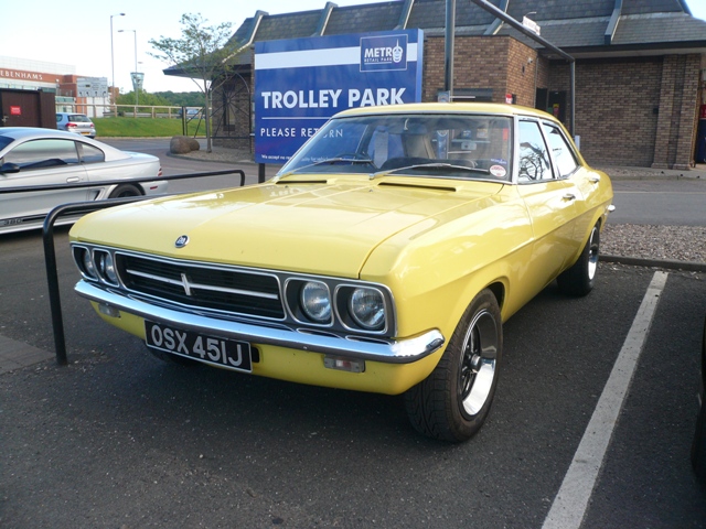 Pistonheads Metrocentre - The image features a vibrant yellow vintage car parked in a parking space. The car is positioned in front of a store, highlighting the store's signage that includes "Trinlley" in white lettering against a blue background. The setting appears to be a parking lot, with some brick buildings and smaller cars visible in the background. The car is equipped with the license plate reading "OSX 451J", giving it a distinctive and unique character.