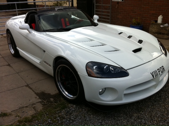 Roll on summer - Page 1 - Vipers - PistonHeads - The image showcases a white convertible sports car, presented in a profile view. The car is parked on a gravel drive next to brick steps, affording a glimpse of the building behind it. The car's design features a prominent air intake in the hood and sleek, shiny detailing. The wheels are adorned with red and black accents, adding a contrasting touch to the white body of the vehicle. The overall setting gives the impression of a quiet and serene day.