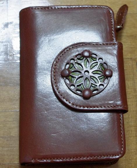 The image showcases a closed, brown leather wallet affixed with a decorative circular metal emblem and four round studs arranged in a square pattern around its midsection. This wallet features a flap with a vertical stitch that secures it when closed. On the top of the flap, two rounded metal studs are visible, suggesting a hinge mechanism. The wallet is placed on a flat surface, and the pattern of the surface's texture can be observed beneath it. The style of the image is straightforward, with no visible text or additional elements, keeping the focus on the wallet itself.