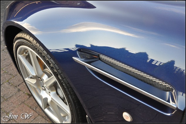 Just had my V8V detailed - just had to share - Page 1 - Aston Martin - PistonHeads - The image showcases a close-up view of the front grill of a black sports car. The car appears to be parked on a brick pavement under a clear blue sky. The grill has a distinctive, sleek design, featuring a silver detail in the center. Another distinguishing feature is a small round silver emblem affixed to the front end of the car. The lighting suggests it might be late afternoon, given the long shadows cast by the car onto the ground. In the bottom right corner, there is a signature that reads "Jim W". The image is taken from a slightly elevated angle, likely by someone standing beside the parked car, capturing the meticulous details of the vehicle's design.