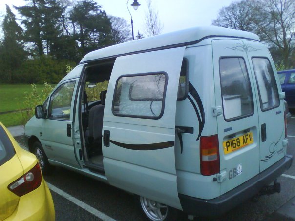 Pistonheads Difference - The image is a photograph of a light blue van parked in what appears to be a parking space. The van has a side window and a clear glass back door. There's a tag that reads "P66 FYV" displayed prominently on the back of the vehicle. The van has a black stripe design along the side. In the background, there is a glimpse of other parked vehicles. The lighting in the photo suggests either early morning or late afternoon, given the soft, diffuse light.