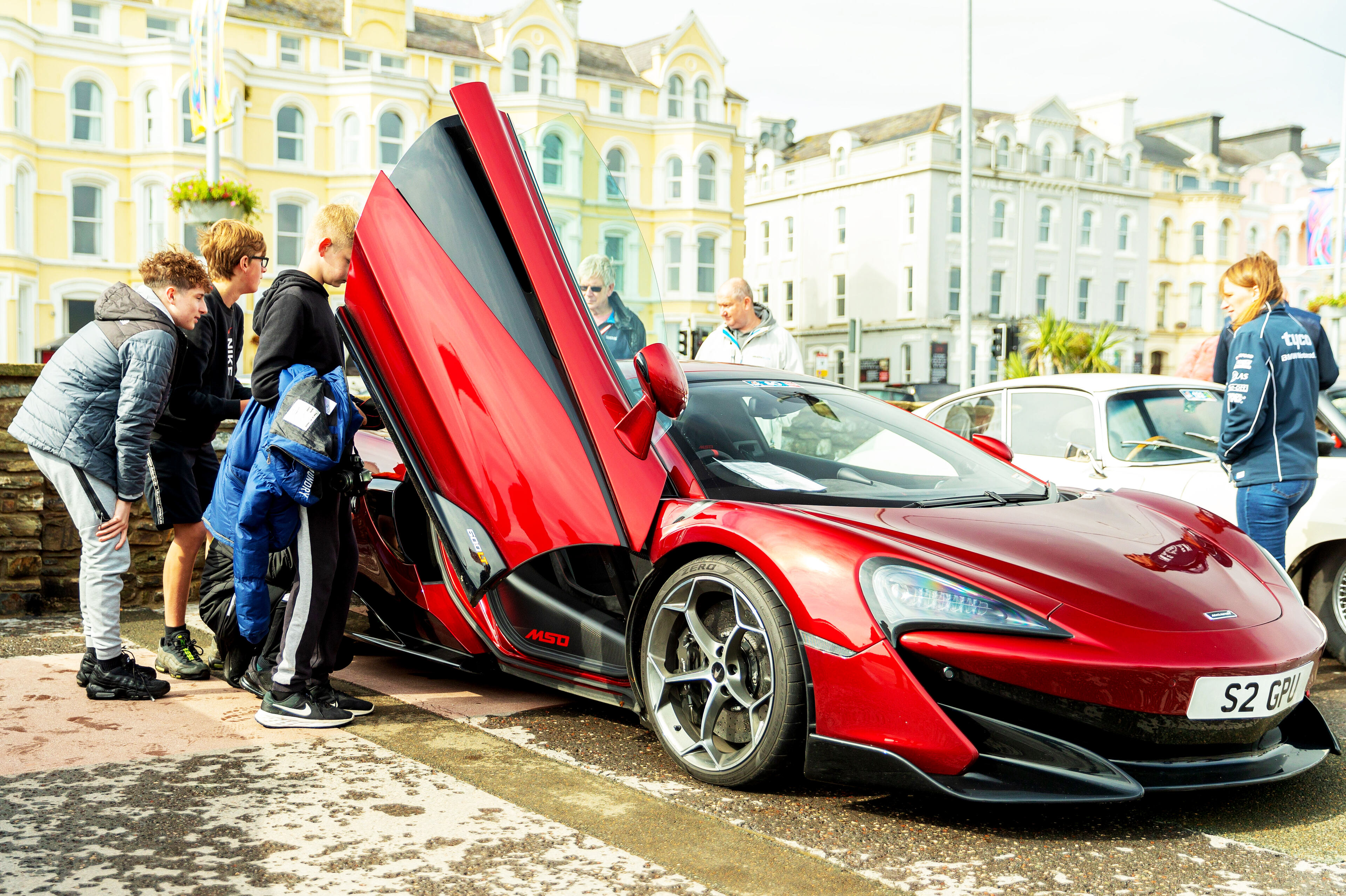 Pistonheads - The image showcases a red McLaren sports car parked in front of a stone building. A group of people is gathered around the car, examining its hood and interior. There are individuals standing at various angles around the vehicle, creating an impression of curiosity and interest. In the background, other vehicles are visible, adding context to the location being a parking lot or a roadside stop where people gather for different reasons. The car's sleek design is accentuated by its vibrant red color, making it a focal point in the image.