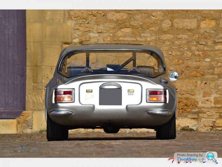 Early TVR Pictures - Page 36 - Classics - PistonHeads - In the image, an old, single-seater GT sports car is parked against a stone wall. The car's chrome bumpers and other details suggest it's a model from the mid-20th century. The background features a stone wall with a slight overlap, a purple door, and a yellowish wall. The focus of the image is on the car, which appears to be front of range rovers. The overall scene gives off a vintage vibe.