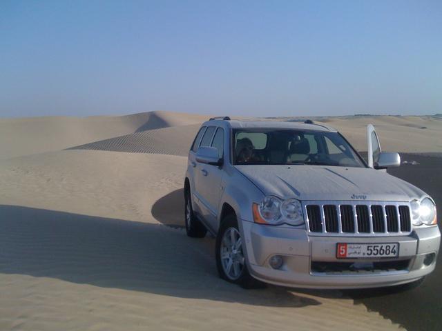 Never off roaded.... - Page 1 - Middle East - PistonHeads - The image captures a striking contrast between the rugged, sandy terrain and the modern, sleek automobile. A silver Jeep, branded with the word "Jeep" prominently displayed on its grille, is parked in the middle of the desert, its black and white license plate reading "5564". The vehicle's presence on this sandy landscape, emphasized by its large size and shiny exterior, creates a juxtaposition of human-made order against the disorderly nature of the desert. The vast, flat expanse of light brown sand meets the deep blue sky at the horizon, with no pronounced features or landmarks visible in the background, further enhancing the sense of isolation and adventure. The Jeep, with its spare tire mounted on the back, appears ready to traverse the challenging terrain, suggesting its owner is prepared for off-road adventures.