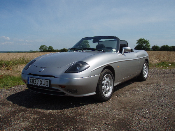 Pistonheads - The image captures a sleek, silver two-door sports car parked on a gravel driveway. It's under the open blue sky, with a field littered with trees in the background. The car has a distinctive flat rear window and is situated on a paved area which appears to be a part of a residential drive. The lack of other vehicles and the tranquil setting suggest it might be during the early morning or late afternoon hours.