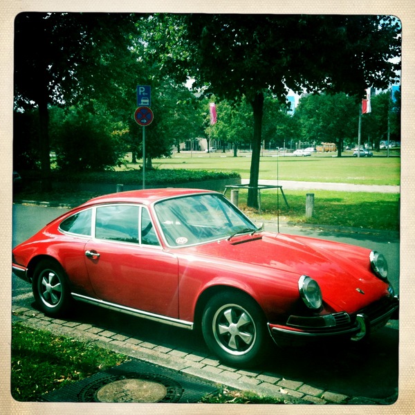 Pistonheads - In the foreground, a striking red classic Porsche 911 Sport Classic is parked on a brick paved area. The car's glossy finish mirrors the surrounding scene. In the background, a park-like setting includes lush greenery, mature trees, and a serene pond. The overall scene is further enriched by the presence of a few people enjoying the park and a majestic white swan gliding gracefully across the pond.