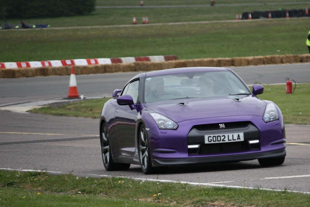 Day Service Dads Pistonheads Sunday - The image showcases a vibrant purple sports car in motion, driving past a red and white striped barrel barrier on a track. The car's license plate reads "God2 Llla". Behind the car, the track curves into the distance, flanked by fields and a line of trees, indicating a rural racing setting. The sky is overcast, casting a soft light over the scene. In the background, there's a person standing at the edge of the track, possibly a marshal or a fellow participant observing the race.