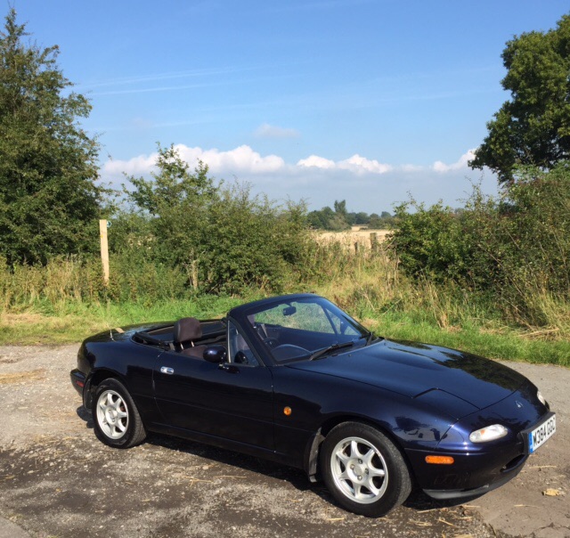 Uh oh I'm going to look at my (potential) 3rd MX5 tonight... - Page 1 - Mazda MX5/Eunos/Miata - PistonHeads - This image shows a black sports car parked on a paved surface. The car features a distinctive convertible top, which appears to be integrated into the design of the vehicle. The vehicle has alloy wheels, a sloping front windscreen, and is positioned at an angle to the camera. The car is situated on a patch of asphalt, with a hedge and green foliage nearby. The photo seems to have been taken during the day under clear skies.