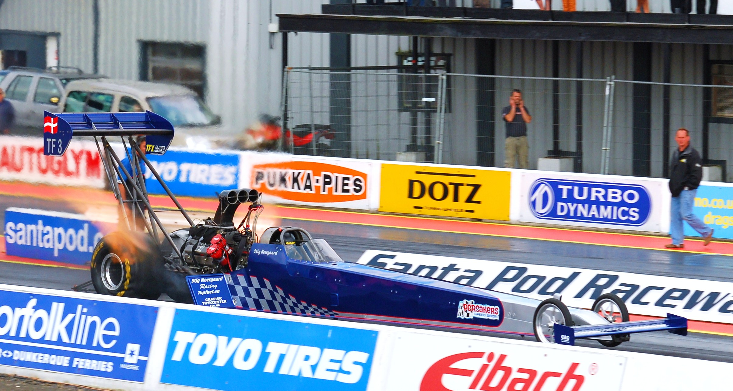 Event Pistonheads Main - The image captures an exciting moment at a car racing event. A racing car is prominently featured in the foreground, zooming past the camera against a contrasting backdrop of blue and white advertisement barriers. There are several other cars and advertising signs in the background, highlighting the competitive and commercial nature of the event. Fans and staff can be seen interspersed amongst the barriers, adding a vibrant atmosphere to the scene.