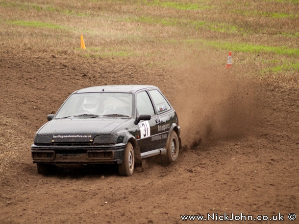 Pistonheads - This image showcases a dynamic scene of a black sports car in action. The car, which is the main focus of the image, is careening on a dirt track. The vehicle has kicked up a significant amount of dust, as evidenced by the plume of dust that is visible in the air above the car. The setting is outdoors, the backdrop is green grass, adding a natural element to the otherwise intense, adrenaline-pumping scene.

The car appears to be a smaller model, with its driver in focus. The driver is actively engaged in maneuvering the car, contributing to the overall sense of motion in the image. The dust kicked up by the car partially obscures the figure of the driver, adding an extra layer of intrigue to this captivating scene.

This image encapsulates the thrilling and energetic spirit of motorsports. The dusted car racing along a dirt road conveys the speed and power at the heart of such racing events. The photographer's perspective, capturing the movement of the car and the dynamic interaction between the car and its environment, presents an exciting snapshot of this racing event.