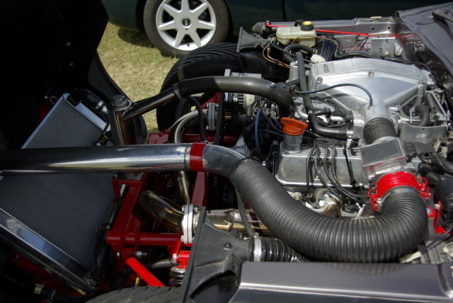 Finished Pistonheads Refurb - The image captures a detailed view of a car's engine compartment. Various components of the engine, including the cylinder head, valves, and other mechanical parts, are visible. Notably, a red-colored pipe runs beside the engine, which might be an exhaust or air intake system. The wires crisscrossing the engine suggest various theatrical or automotive effects using lighting and smoke. The background, described as "grassy", provides a contrast to the mechanical precision of the engine.