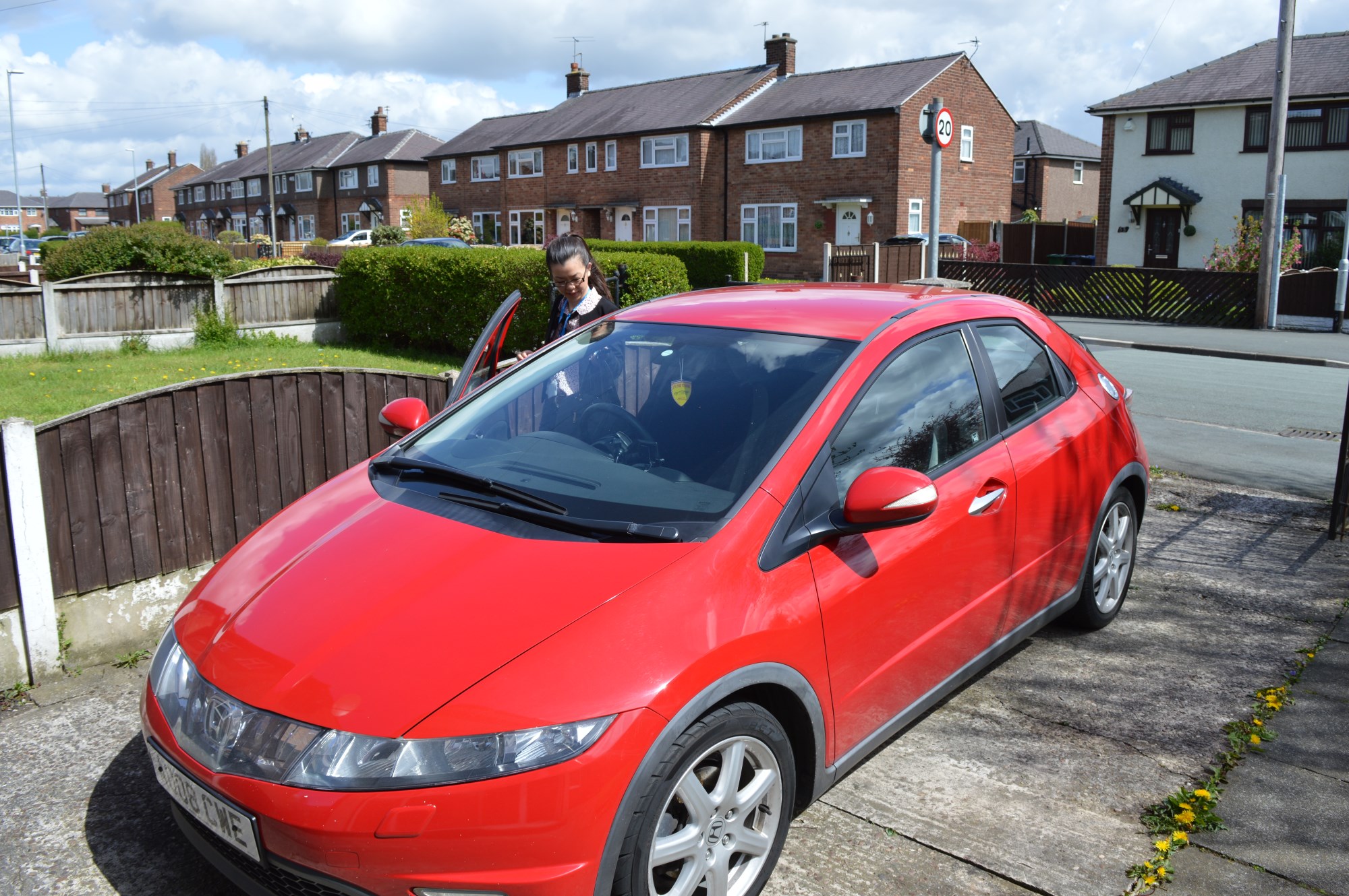 How Do Civics Handle High Miles? - Page 1 - Honda - PistonHeads - The image shows a red hatchback parked on a concrete driveway. To the left of the car, a person appears to be attending to the vehicle's interior by possibly reaching into or touching the car door. The setting suggests a suburban or semirural area, indicated by the presence of a fence at the end of the driveway and a car parked behind the featured vehicle. The sky is visible in the background, and the time of day seems to be during daylight hours. There are no visible texts or distinctive brands in the image.