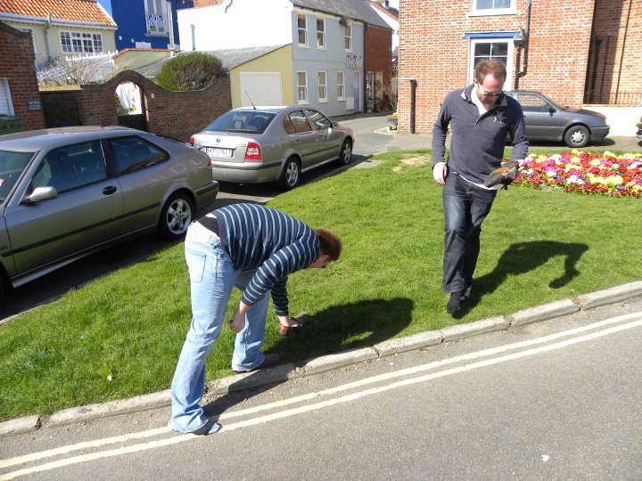 Funniest Pistonheads Quote - The image captures a street scene with a pair of individuals engaged in the act of bending over to reach something on the ground. One person is wearing a blue shirt and blue jeans while the other sports a black shirt and black pants. They are both focused on the task at hand. The setting includes parked cars that line the street and a lush green grassy curb. Further back, a brick building is visible, adding a sense of depth to the image. The atmosphere seems calm and unhurried, as if a leisurely walk has led to a moment of careful inspection or retrieval.