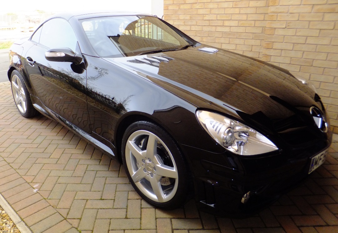 Mercedes SLK 55 AMG - Page 2 - Readers' Cars - PistonHeads - The image shows a sleek black convertible car parked on a cobblestone driveway, with the hood reflected in the adjacent brick wall. The car's glossy finish reflects the surrounding area, enhancing the gleam of its bodywork. The vehicle's round wheels and white rims are prominently displayed. A notable feature is the large front grill, which adds to the car's aggressive and stylish design. The setting appears peaceful, hinting at private property rather than a public parking area.