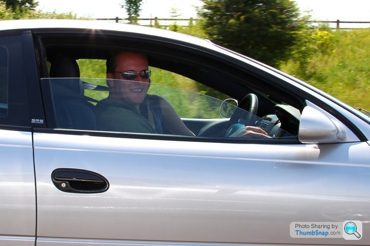 Pistonheads Monaro Vxr - The image depicts a man driving a silver car under a bright sky, suggesting good weather. The man is wearing sunglasses and a green shirt, and he's looking at the camera with a smile. The interior of the car is visible through the windows, and the mirror indicates that he's on the road. The car's sleek design is apparent on the side and front, and the road is lined with trees or bushes, adding to the natural ambiance of the scene.