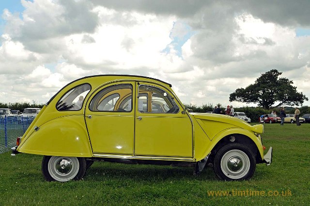 2CVGB Registers Day 2012  - Page 1 - Photography & Video - PistonHeads - The image captures a vibrant scene set in the UK. Dominating the foreground is an antique yellow Volkswagen Beetle, its hood is open, revealing an engine that promises an amusing test drive. This Beetle is not shown as a tourist attraction but as part of an exciting event. Surrounding the Beetle, the grass glistens under the presence of scattered cars and people, painting a casual and lively atmosphere. The backdrop is a serene mix of an ancient tree and a clear blue sky, adding a touch of nature's beauty to the image. The overall setting seems to be a car show, where car enthusiasts gather to celebrate the charm of vintage vehicles.