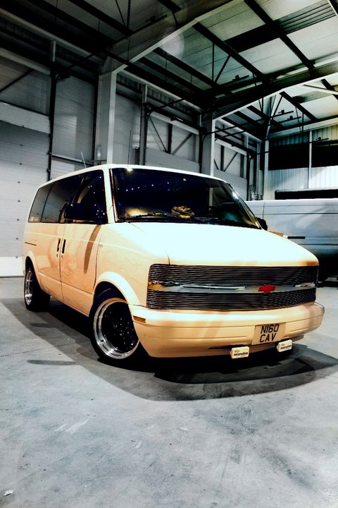 my show van  - Page 1 - Yank Motors - PistonHeads - This is a garage-style photograph focused on the front of a golden or bronze HHR van, with the vehicle positioned centrally in the image. The van is a hatchback style with tinted windows and a distinctive front grille. There is either a sunroof or an additional panel of glass, and no visible wheel covers are shown. The background features a high overhead door and other industrial structures that suggest an indoor setting, likely a hangar or workshop. The lighting in the image is soft, contributing to the vehicle's metallic sheen.