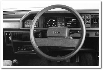 Pistonheads Metro - The image presents an interior view of a vehicle's dashboard, centered around a steering wheel. The steering wheel, which is the main focal point of the image, is made of beige leather, appearing old. The dashboard houses a range of gauges and controls, including a clock on the left side and a radio system, all under a hood that features an open engine compartment in the background. The photograph is in black and white, adding a timeless quality to the scene.