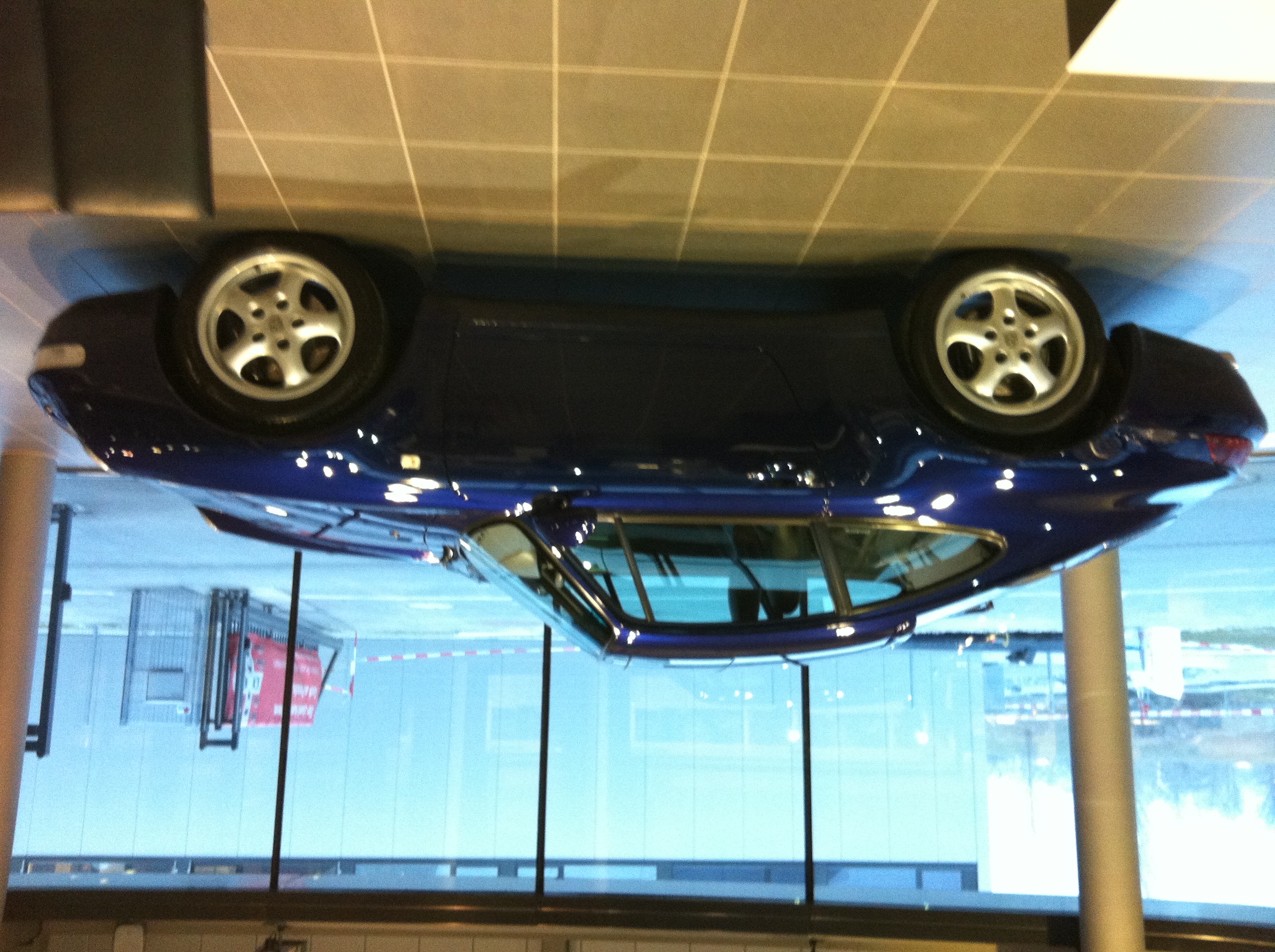 Pistonheads - The image shows a vibrant blue car displayed on a white stand, giving the impression of the car being suspended in mid-air. The car is an exotic sports model, characterized by its sleek design and numerous wheels. The setting appears to be indoors, with the ceiling visible above the display. There are no people present in the image.